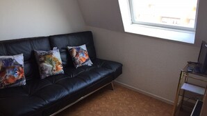 A black leather sofa with colorful pillows, a wooden parquet floor, and a window with a view of buildings.