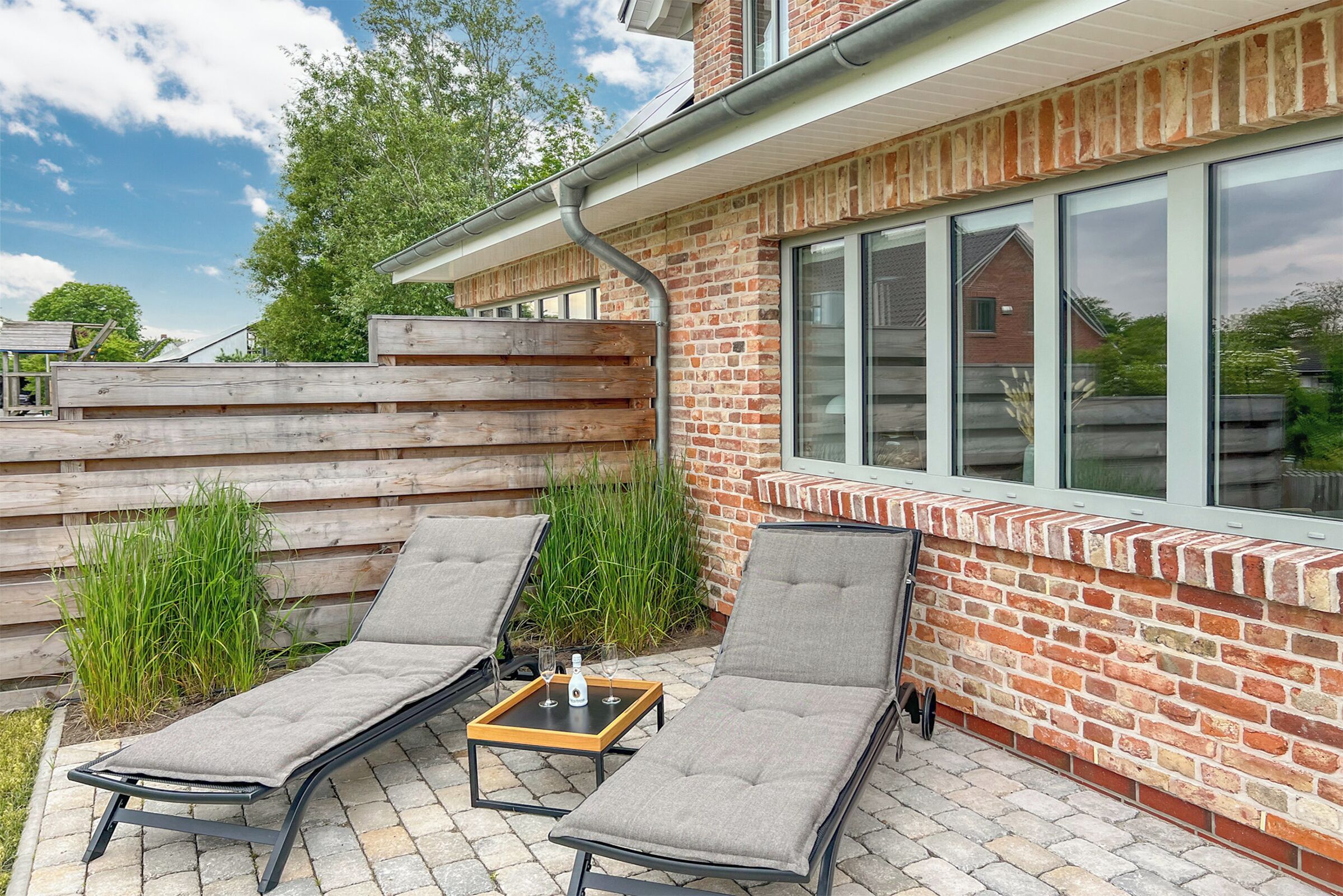 A patio with two lounge chairs and a small table, a brick wall, and a window overlooking a garden.