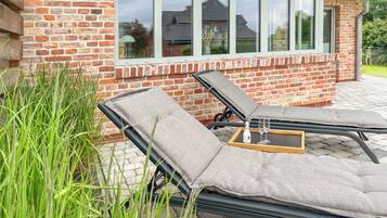 A brick house with a patio area featuring two lounge chairs and a table with a glass.