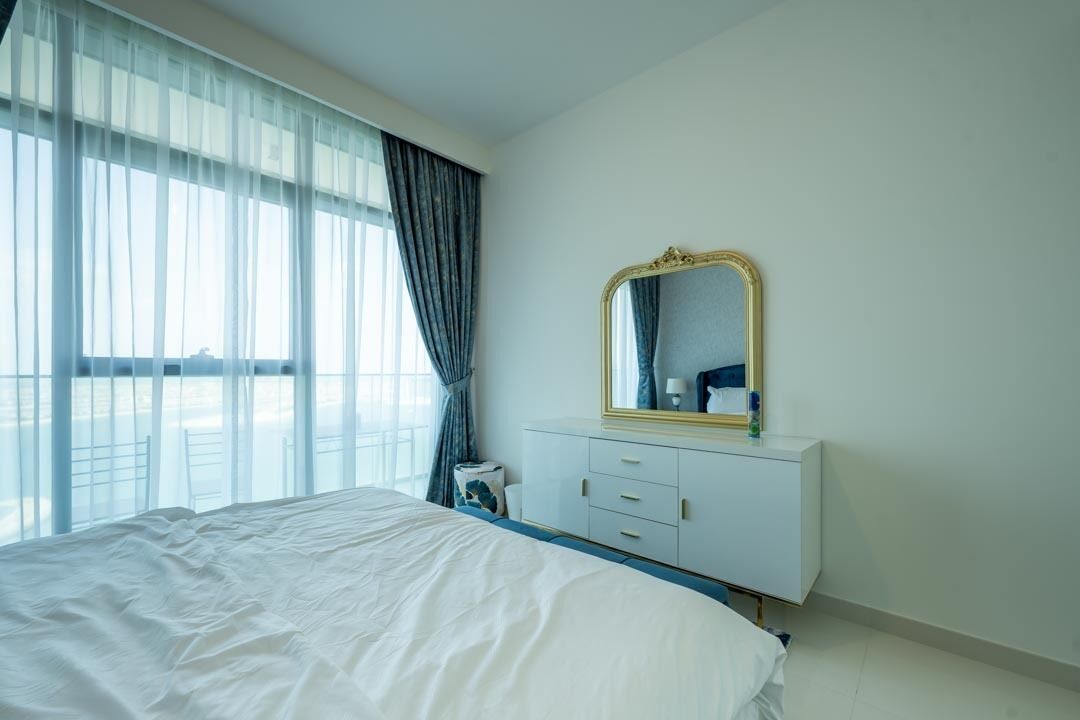 Une chambre d’hôtel avec un grand lit, une commode blanche avec un miroir et une vue dégagée sur l’extérieur grâce à de grandes fenêtres.