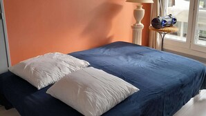 A bed with a dark blue cover and two white pillows in a room with an orange wall and a lamp on a side table.