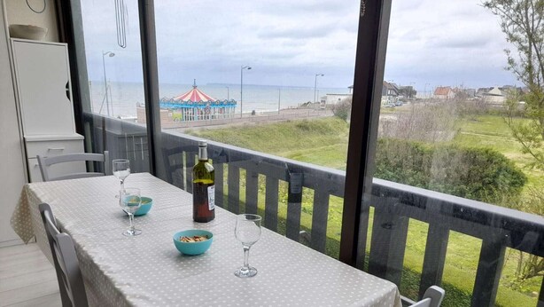 A dining table with a polka dot tablecloth, two bowls, a bottle of wine, and two wine glasses on a balcony overlooking a beach with a carousel.