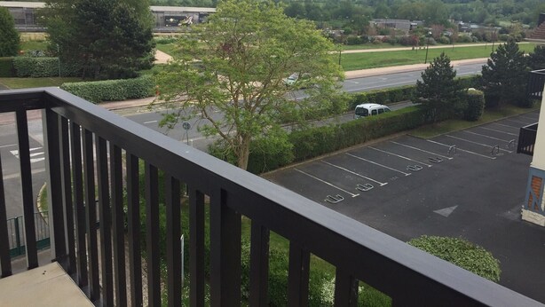 Une vue depuis un balcon donnant sur un parking, avec un arbre au premier plan et une route en arrière-plan.