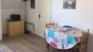 A dining area with a table covered in a patterned tablecloth, a chair, a floor lamp, a television on a wooden cabinet, and a door with a picture on the wall.