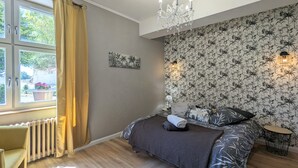 A bedroom with a bed, a radiator, a window with curtains, a chandelier, and a wall with a floral wallpaper.