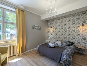 A bedroom with a bed, a radiator, a window with curtains, a chandelier, and a wall with a floral wallpaper.