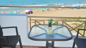 Un balcón con una mesa y sillas de vidrio, con vistas a una playa con sombrillas y gente.