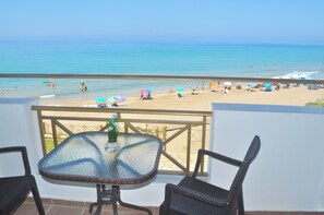 Un balcon avec une table en verre et des chaises en osier, donnant sur une plage parsemée de parasols et animée par la présence de nombreuses personnes.