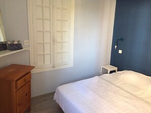 Quarto com cama, cómoda de madeira, janela com persianas brancas e mesa de cabeceira com abajur.