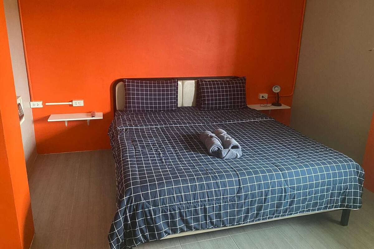 A single bed with a checkered bedspread, a pillow, and a rolled towel on the bed. There is a small bedside table with a lamp, and a power outlet on the wall.