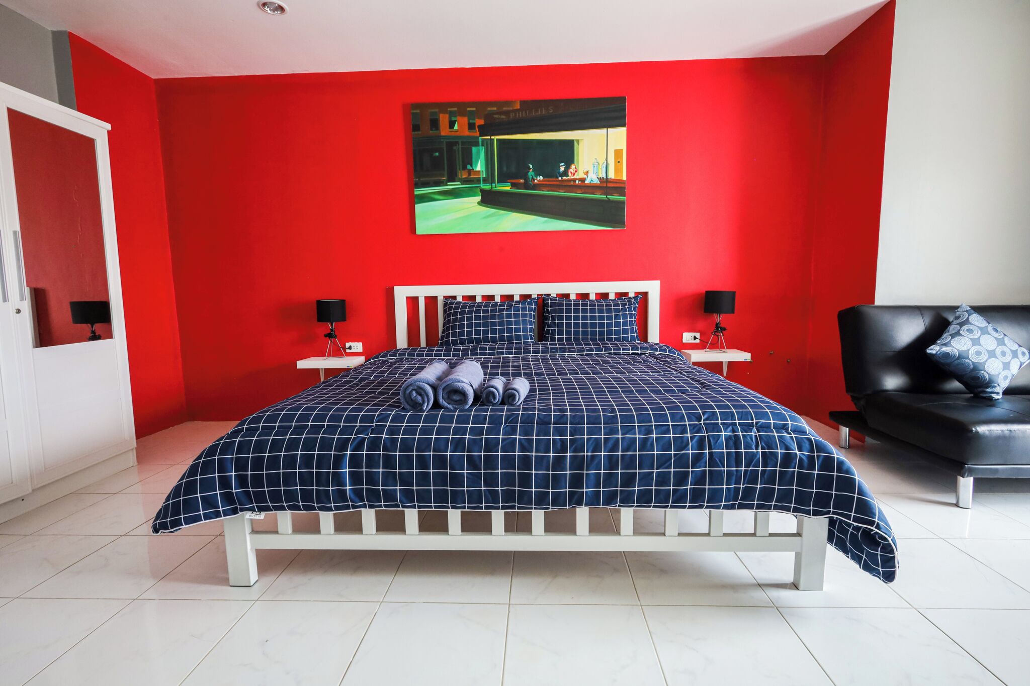 A bedroom with a red wall, a bed with a checkered blue and white duvet, two bedside tables with lamps, and a black leather sofa with a patterned pillow.