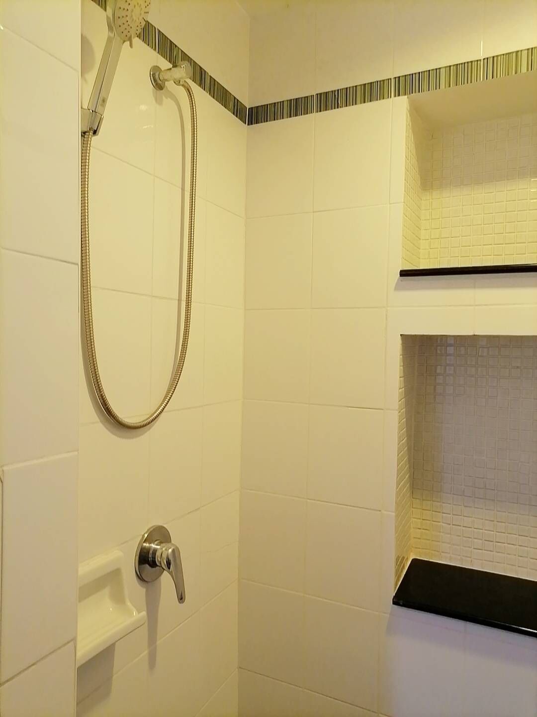 A bathroom with white tiles, a shower with a handheld showerhead, and a black countertop.
