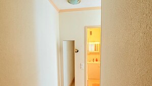 Un estrecho pasillo que conduce a una habitación con puerta, un espejo y un baño pequeño con inodoro y lavamanos.