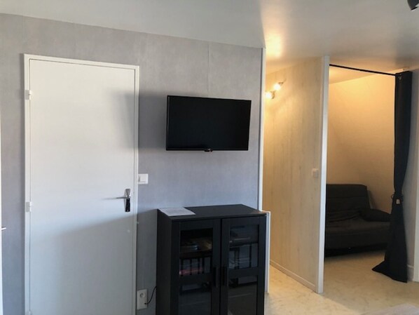 Una habitación con una puerta blanca, un mueble negro con puertas de cristal, un televisor montado y un sofá de color oscuro al fondo.