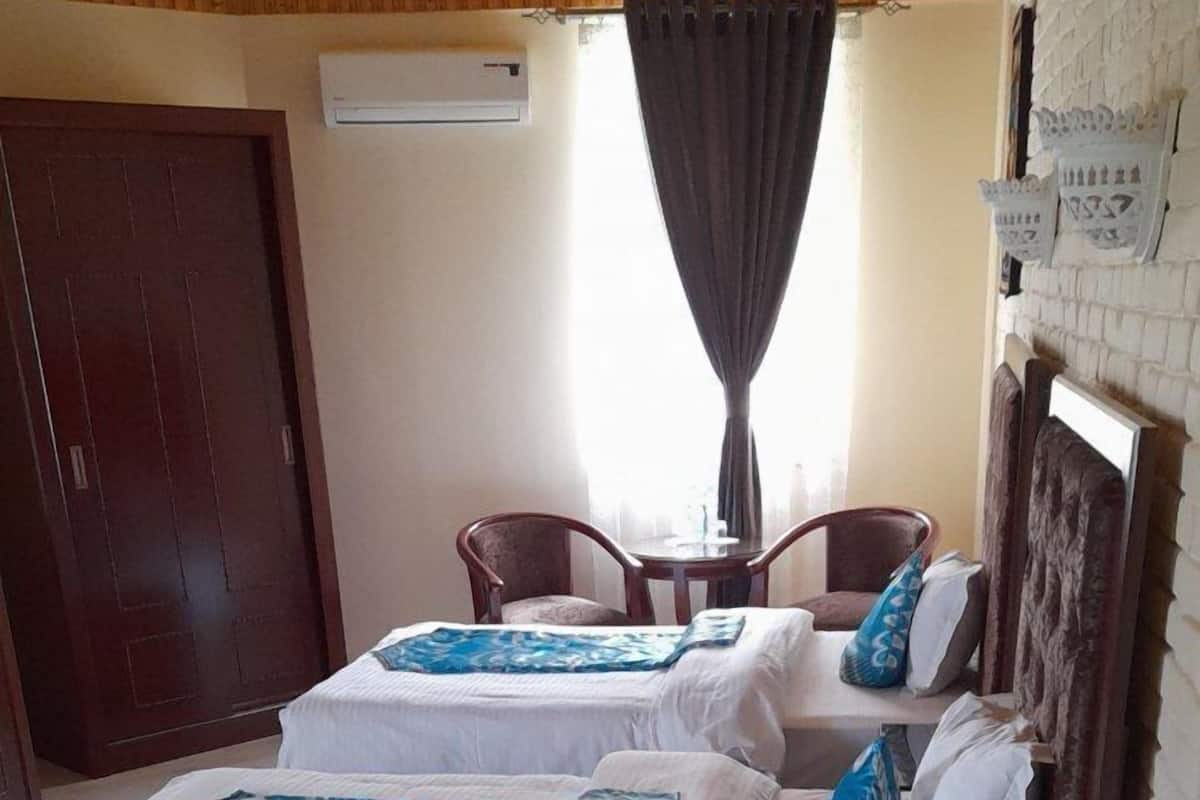 A double bed with white bedding and blue accent pillows, a wooden headboard, a small table with chairs, and a dark wooden door.