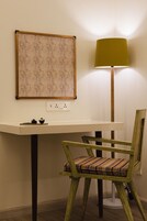 Una habitación con un escritorio blanco, una silla verde con cojín estampado, una lámpara de pie y una obra enmarcadas en la pared.
