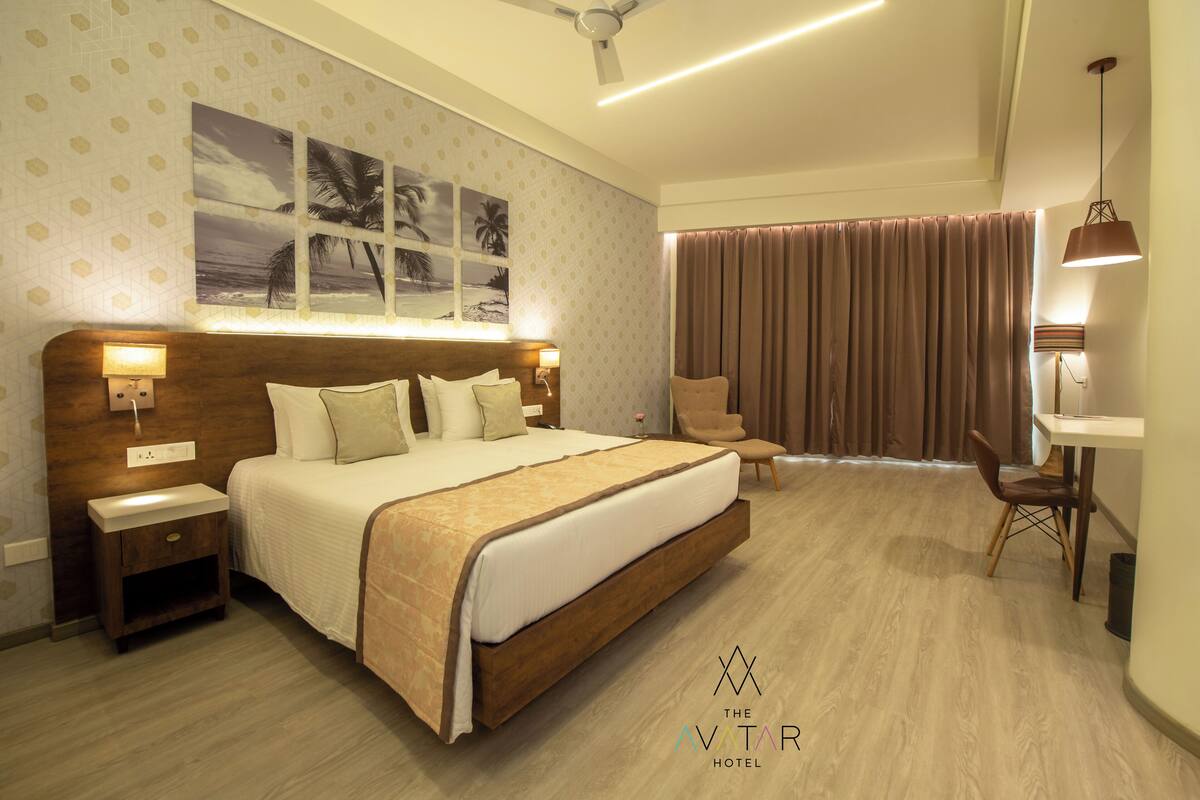 A hotel room with a large bed, two bedside tables, a desk with a chair, a ceiling fan, and a large window with curtains.