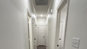 A narrow hallway with white walls and wooden flooring, leading to a door at the end. - Stay Tyler @Royal Hills (Tyler)