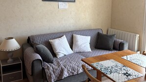 Ein gemütliches Wohnzimmer mit einem grauen Sofa, einem Holztisch mit gemusterten Platzsets, einer Lampe und einem Gemälde einer Küstenlandschaft an der Wand.