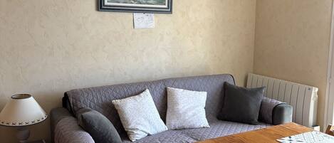Un salón acogedor con un sofá gris, una mesa de madera con manteles individuales estampados, una lámpara y un cuadro de una escena costera en la pared.