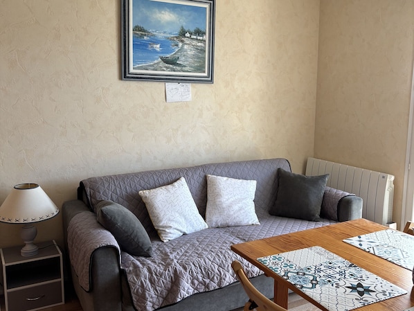 A cozy living room with a grey sofa, a wooden table with patterned placemats, a lamp, and a painting of a coastal scene on the wall.