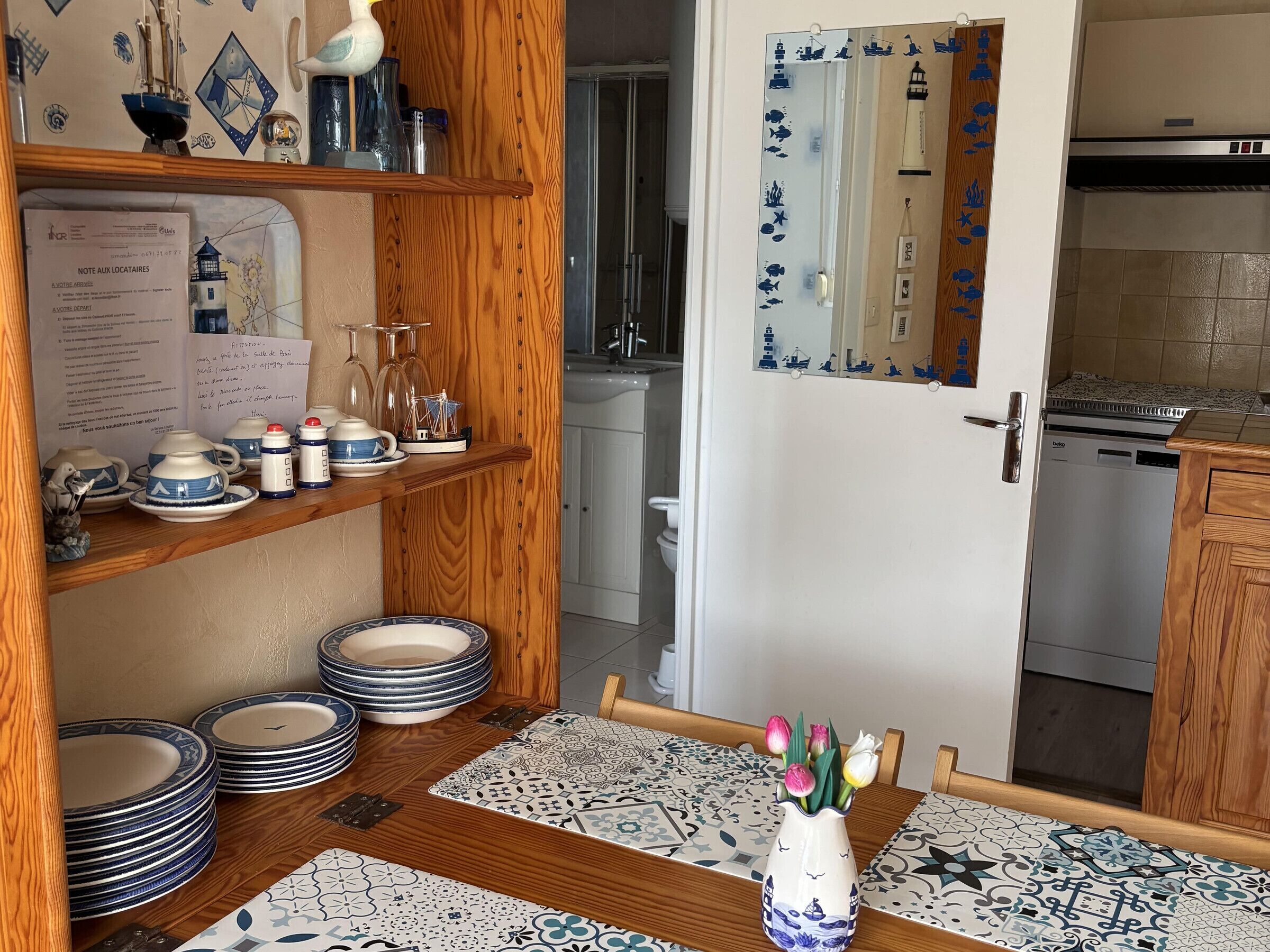 A kitchen with wooden shelves, a dining table with patterned tablecloths, and a vase with flowers.