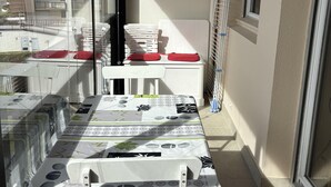 Ein Balkon mit Tisch, bedeckt mit einer gemusterten Tischdecke, weißen Stühlen mit roten Kissen und einem Wäscheständer an der Wand.
