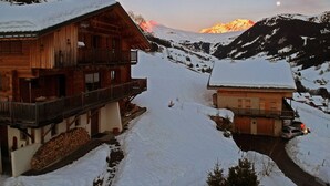 A wooden chalet with a balcony, a snow-covered path, and a mountain in the background.