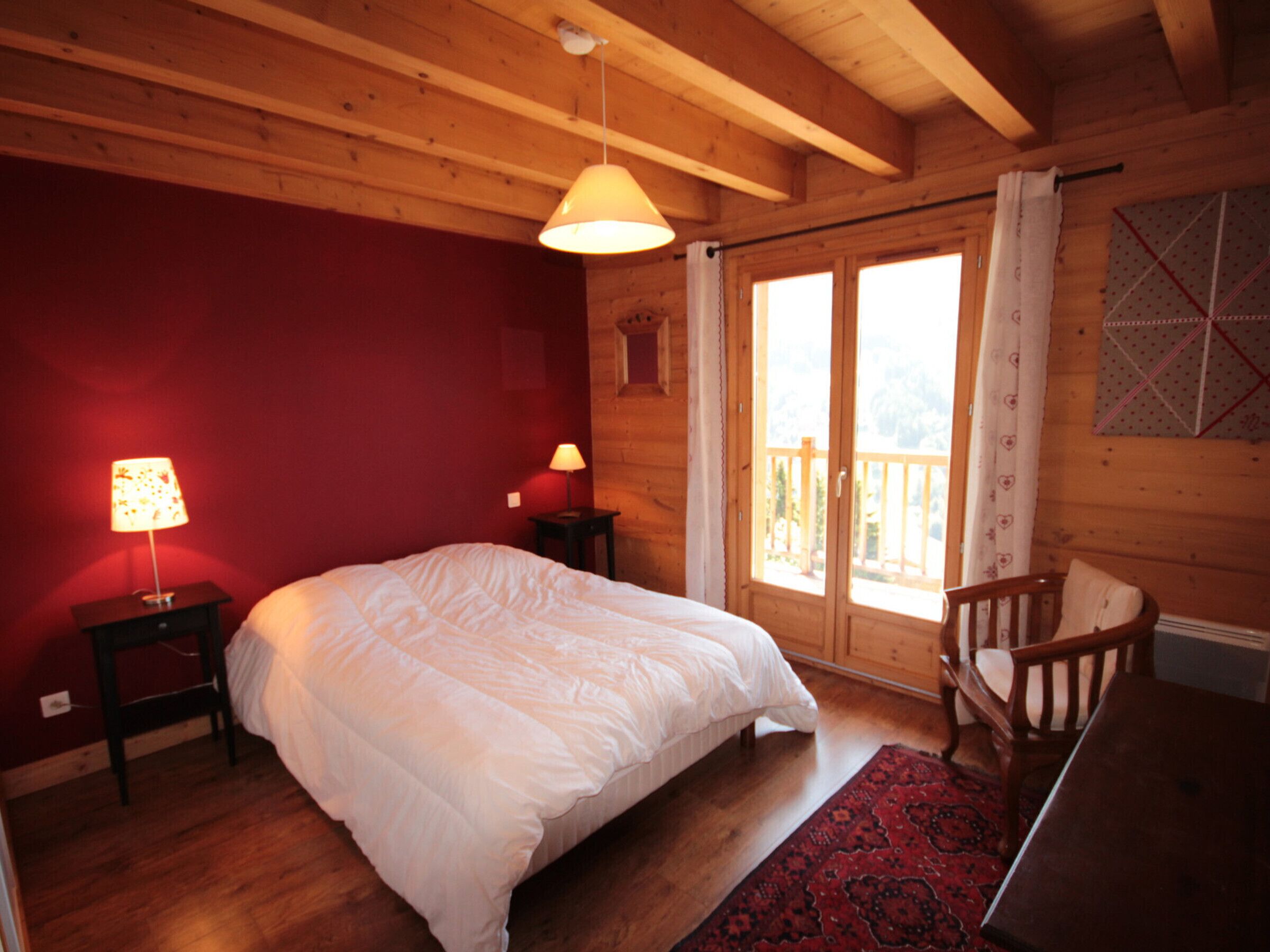 Un dormitorio de cabaña de madera con una cama, dos lámparas de noche, una silla de madera y un ventanal con cortinas.