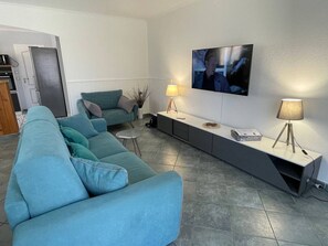 Uma sala de estar moderna com um sofá azul-turquesa, uma TV de tela plana na parede, uma mesinha de cabeceira com abajur, e uma cozinha ao fundo.