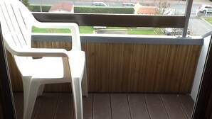 Un balcon avec une chaise en plastique blanche, un plancher en bois et une vue sur un quartier résidentiel.