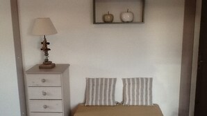 Une petite pièce simple, avec un banc en bois, une commode blanche à tiroirs, une lampe et une étagère sur laquelle reposent deux pommes.