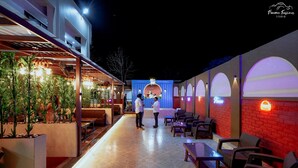 Un área de restaurante al aire libre con un espacio de estar techado, plantas en macetas y un escenario para espectáculos.
