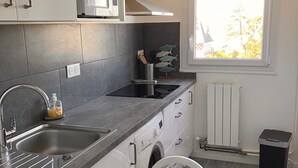 A modern kitchen with a built-in washing machine, a sink, and a window with a view of trees. - 2-room apartment with balcony steps from the beach, WIFI and parking (Villers-sur-Mer)