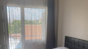 A neatly made bed with white linens in a room with a large window, a dark headboard, and a view of the outside. - Taner Pansiyon (Edirne)
