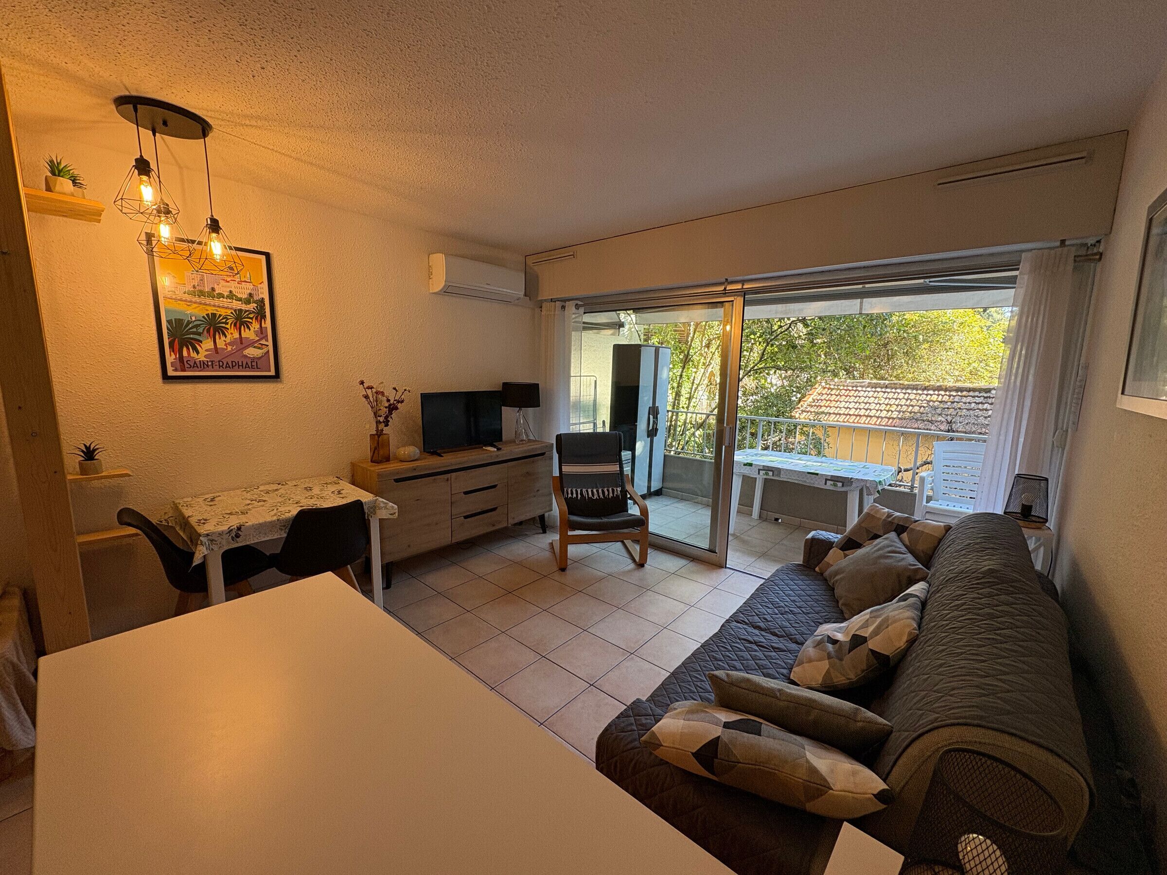 A cozy living area with a sofa, a dining table, and a TV. There's a balcony with a view of trees and a house.