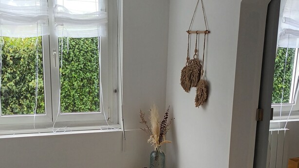 A room with a wooden staircase, a round woven rug, a vase with dried plants, and a wall-mounted macrame ornament. - THE HOLIDAYS (Arques)