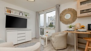A modern living room with a flat-screen TV, a sofa, a dining table with chairs, a bookshelf, and a large window with curtains. - Bright Studio with Balcony | Tranquil Zoliborz (Warszawa)