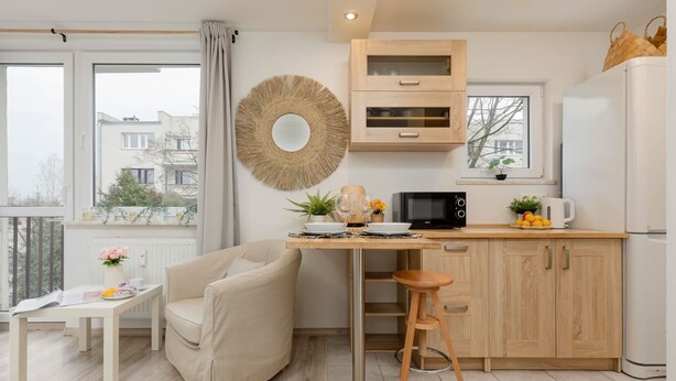 A modern kitchen with a wooden cabinet, a microwave, and a refrigerator. There is a dining table with a chair and a small stool. A large window provides a view of the outside. - Bright Studio with Balcony | Tranquil Zoliborz (Warszawa)