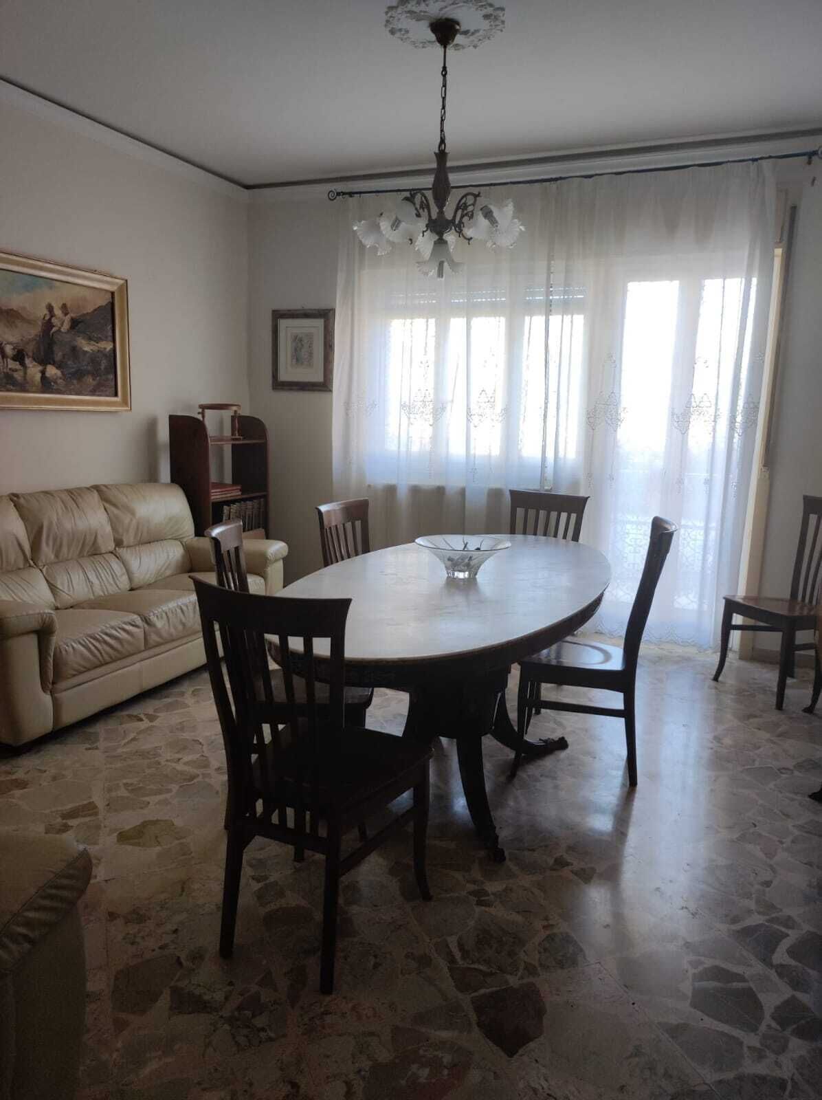 Un comedor con una mesa redonda, seis sillas, una araña, un sofá, una estantería y un cuadro en la pared.