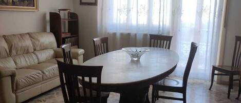 Un comedor con una mesa redonda, seis sillas, una araña, un sofá, una estantería y un cuadro en la pared.