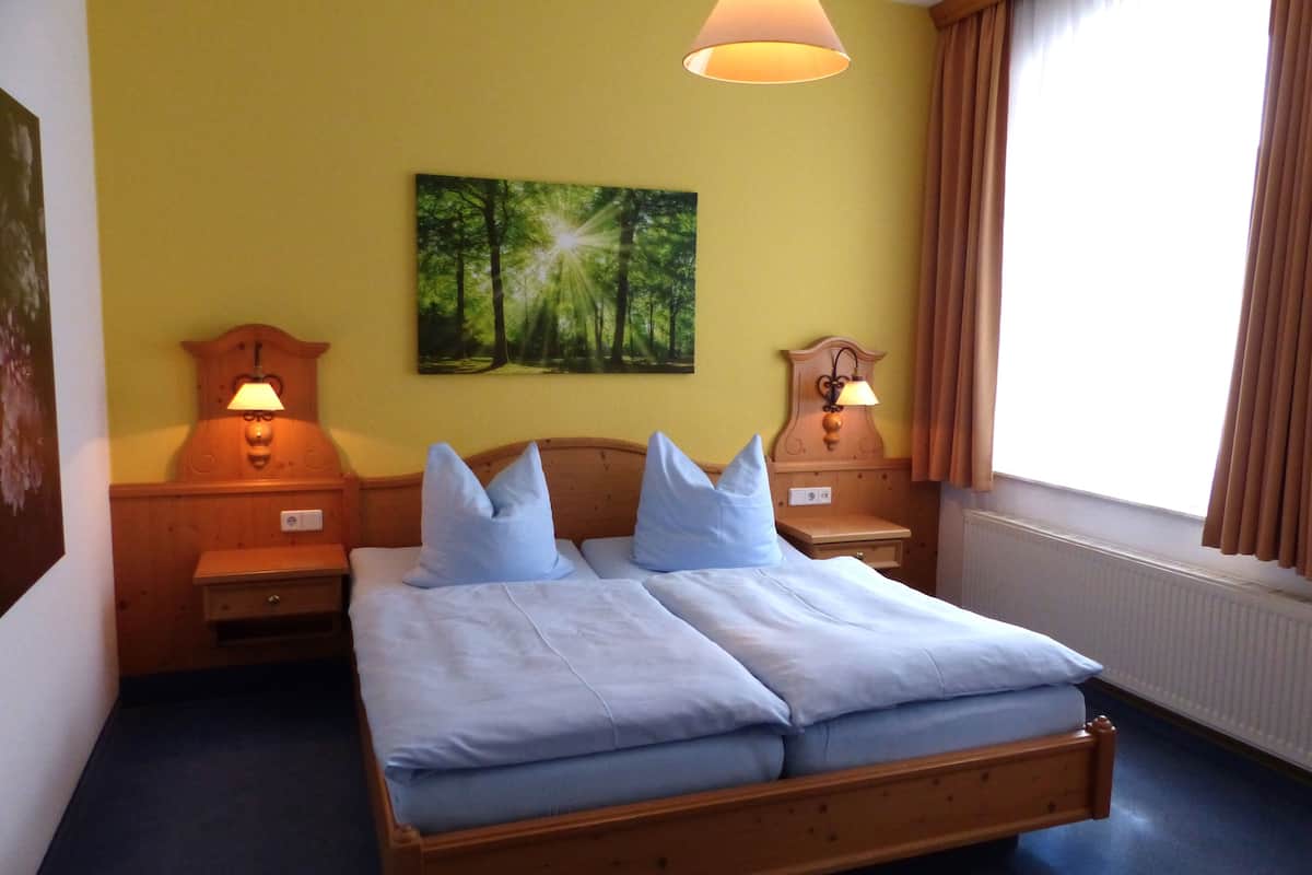 A double bed with light blue bedding, two nightstands with lamps, a wall-mounted painting of a forest, and a window with curtains.