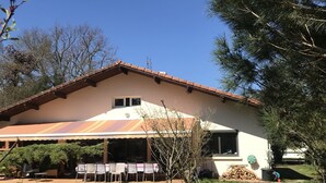 A building with a covered patio area, outdoor seating, and a small tree in the foreground. - Messanges - Landes: Villa Waves & Olas (Messanges)