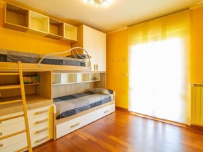 A room with a bunk bed, a desk, and a chair. There are built-in shelves above the bed and a window with curtains.