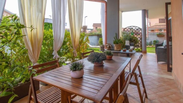Ein Holztisch mit Stühlen und Blumentöpfen auf einer Terrasse, mit Blick auf Grünflächen und Gebäude im Hintergrund.