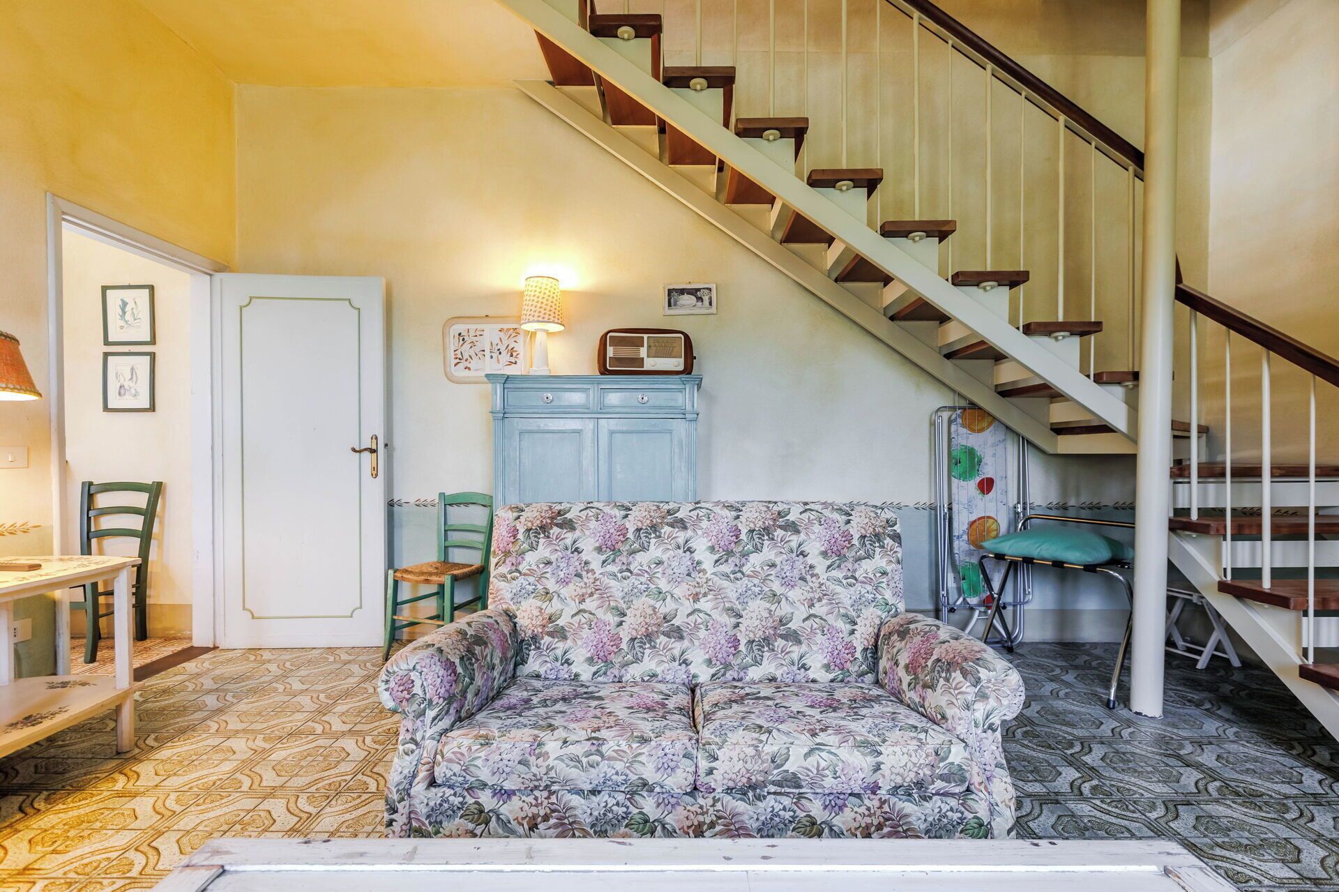 A room with a floral-patterned sofa, a wooden staircase, a vintage radio, and a patterned floor.