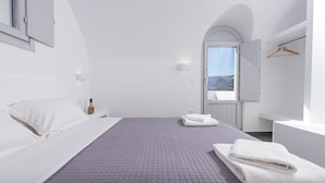 A minimalist bedroom with a large bed, a view through an open door, and a hanging clothes rack. - Golden View Jacuzzi Suites (Santorini)