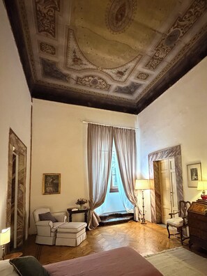 A room with a wooden floor, a bed, a chair, a table with a lamp, and a window with curtains. - Renaissance Palazzo Apartment Decorated with Frescoes Near the Duomo - Sleeps 8 (Firenze)
