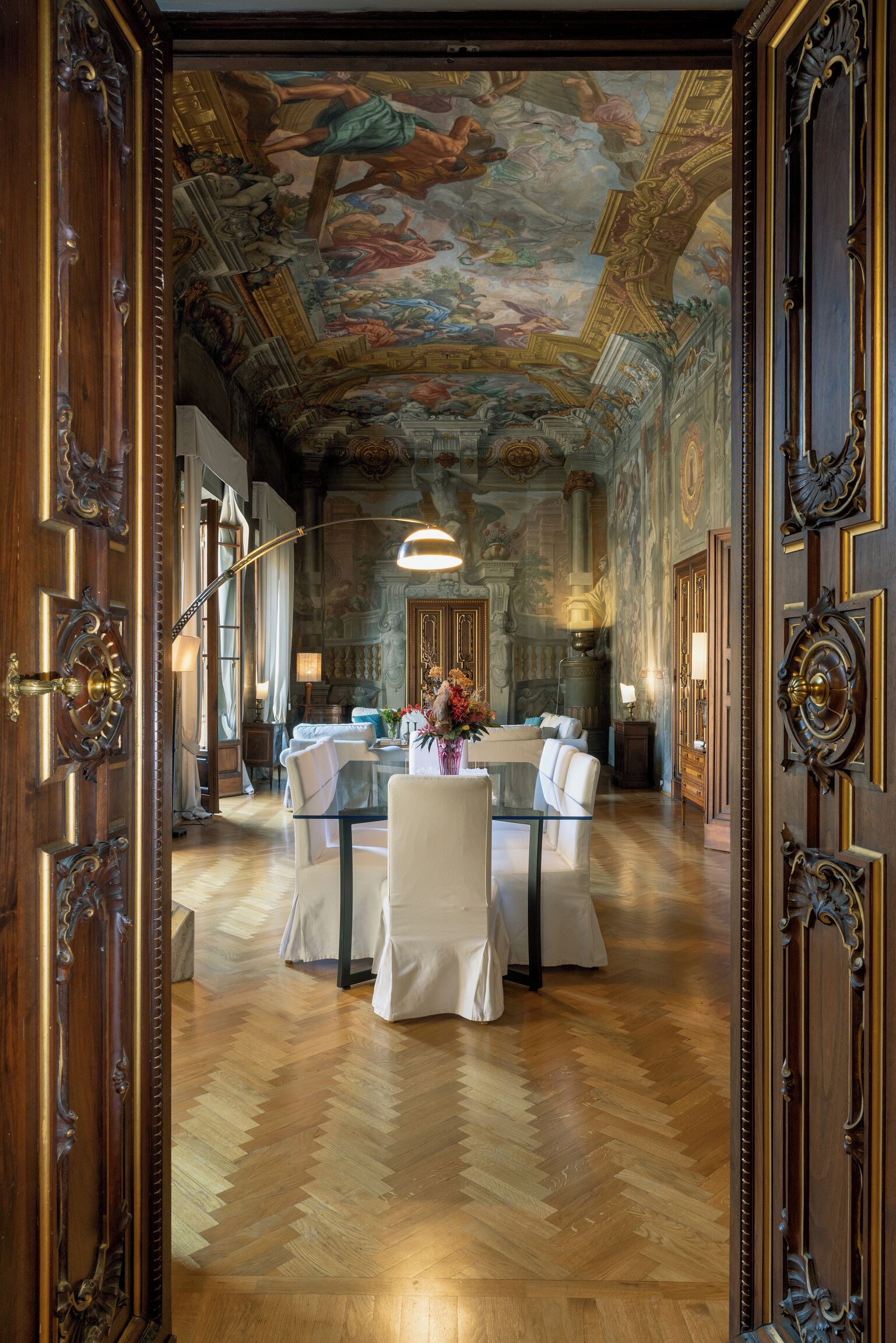 Een ruime kamer met een hoge plafond, versierde houten deuren, een eethoek met een glazen tafel, en een gedetailleerde fresco op het plafond.