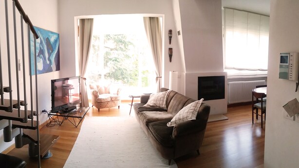 Ein geräumiges Wohnzimmer mit einer Couch, einem Sessel und einem Fernseher. Eine Treppe führt zu einer weiteren Ebene.